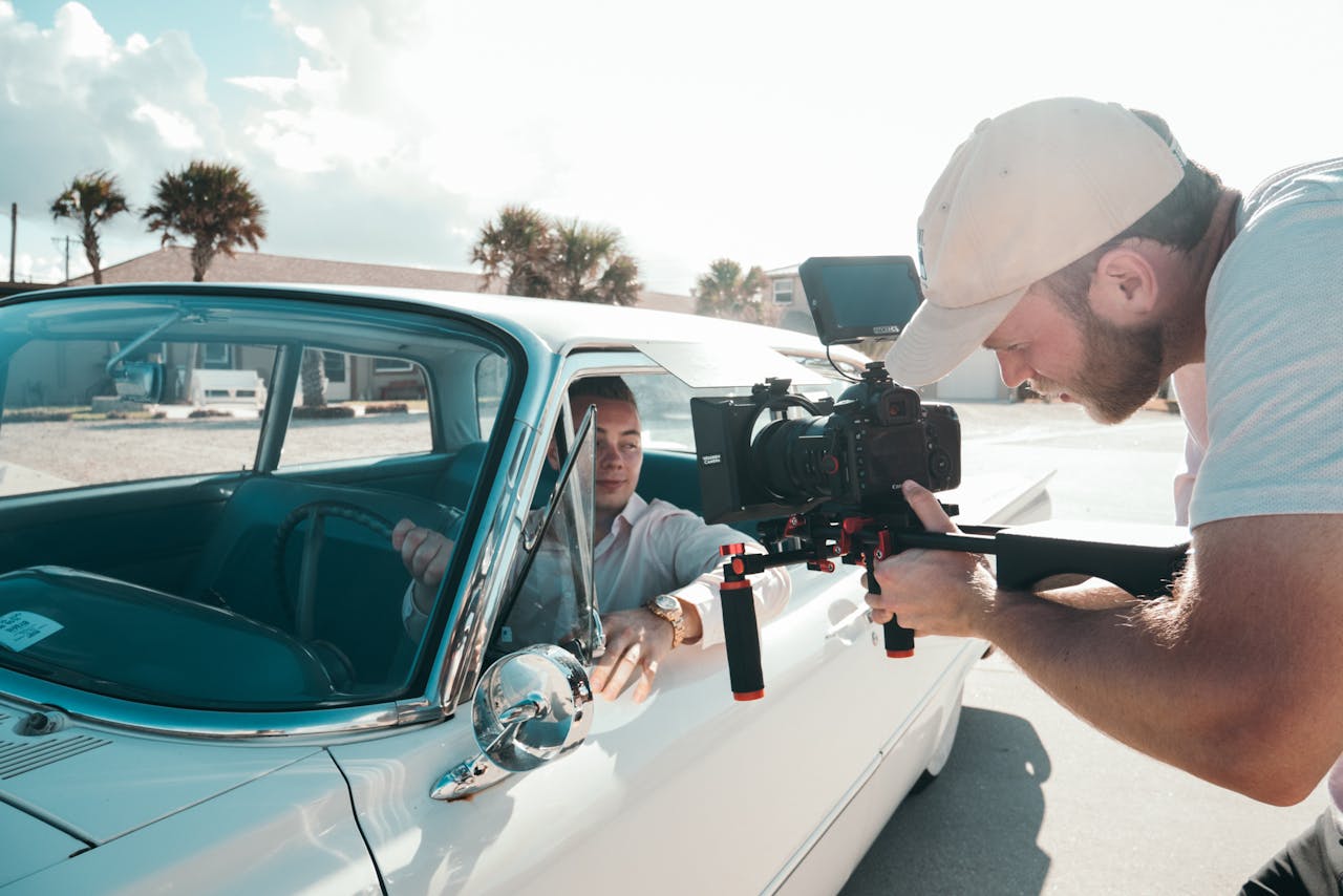 Behind-the-scenes shot of a videographer filming an actor in a classic car during daytime.